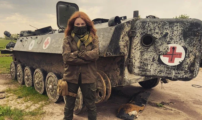 Жінка з рудим волоссям стоїть перед броньованою медичною машиною в полі
