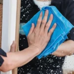 Людина протирає дзеркало синьою ганчіркою, ілюструючи помилки догляду засобом для скла, що руйнує поверхню.