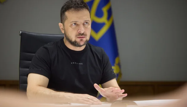 Володимир Зеленський у чорній футболці жестикулює за столом біля прапора України