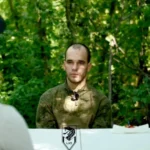 Молодий чоловік у камуфляжі сидить за білим столом у лісі навпроти співрозмовника