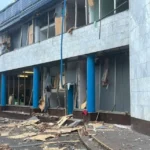 Пошкоджений фасад будівлі з синіми колонами, вибитими вікнами та уламками на тротуарі