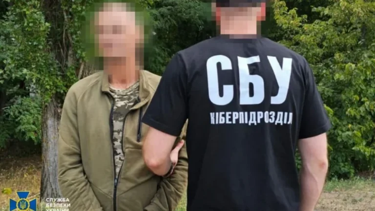 Працівник кіберпідрозділу СБУ тримає затриманого в оливковій куртці серед дерев