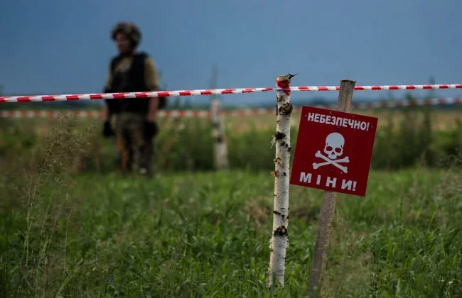 Попереджувальний знак про міни на полі біля солдата та огороджувальної стрічки